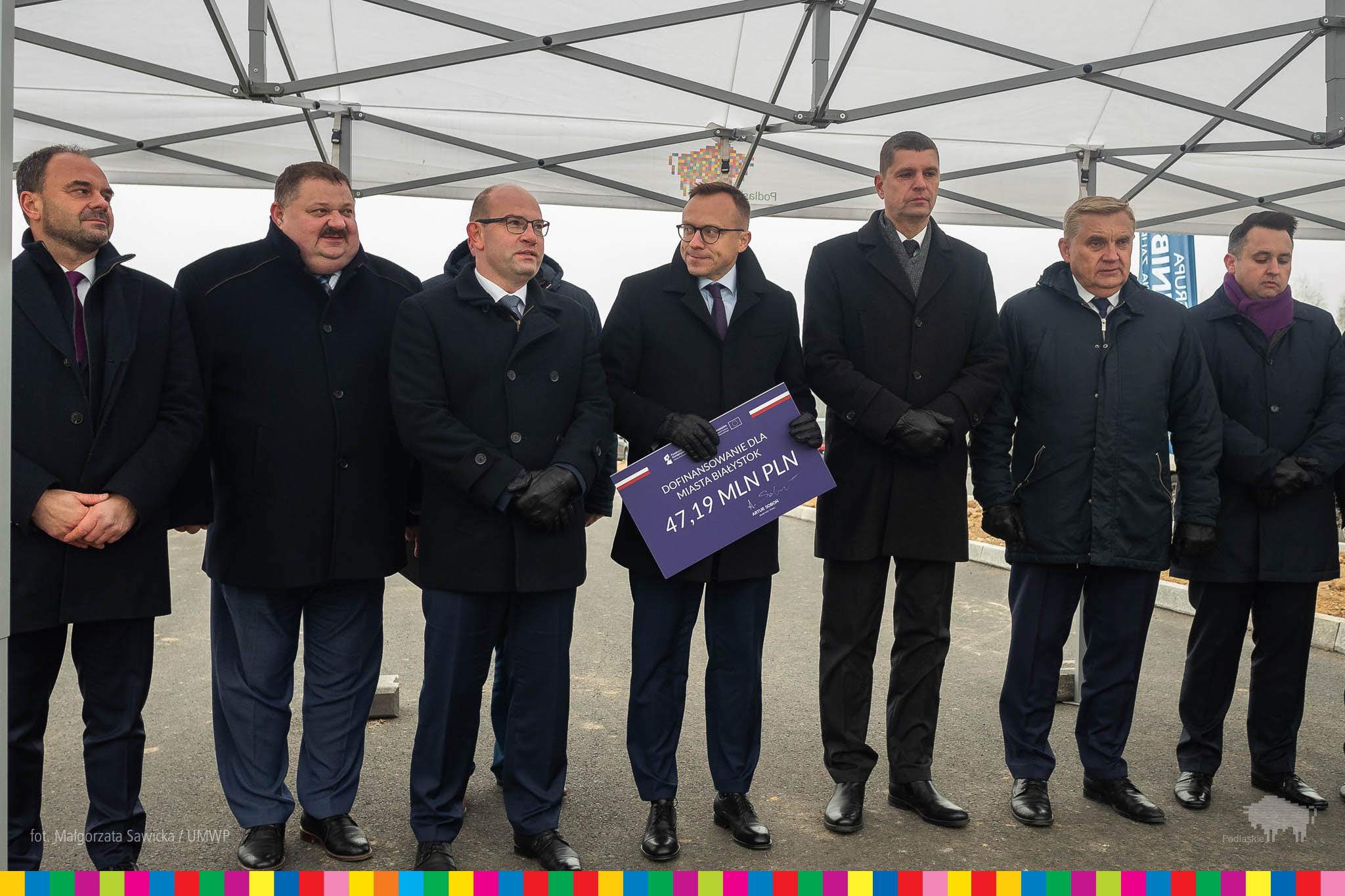 Pod białą wiatą ustawioną na terenie budowy stoi grupa mężczyzn w czarnych garniturach, jeden z nich trzyma czek symbilizujący otrzymane dofinansowanie.